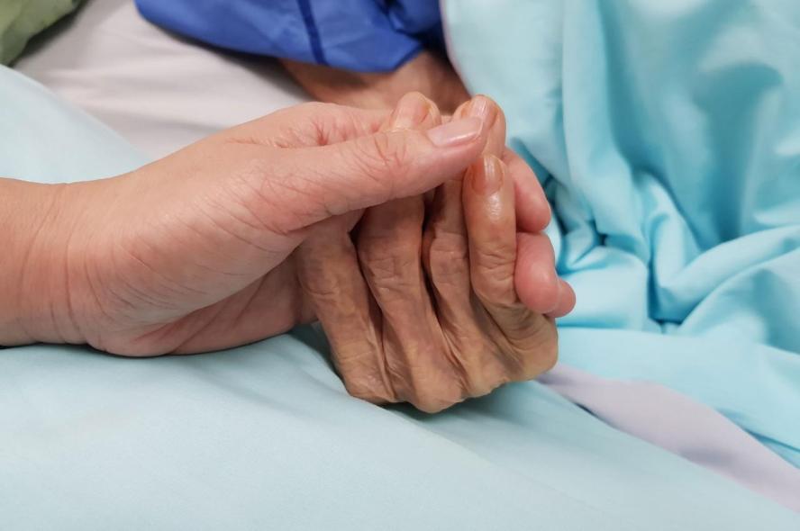 An elderly patient in a hospital bed holds hands with a doctor