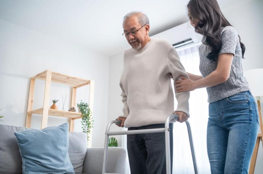 An older man using a walker at home receives help walking from a young woman