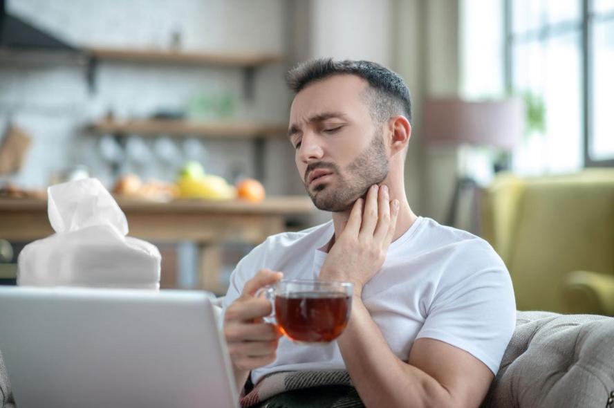 A young man with a sore throat drinks tea on the couch