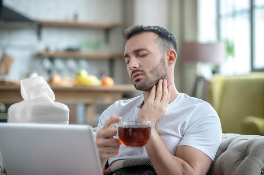 A young man with a sore throat drinks tea  on the couch