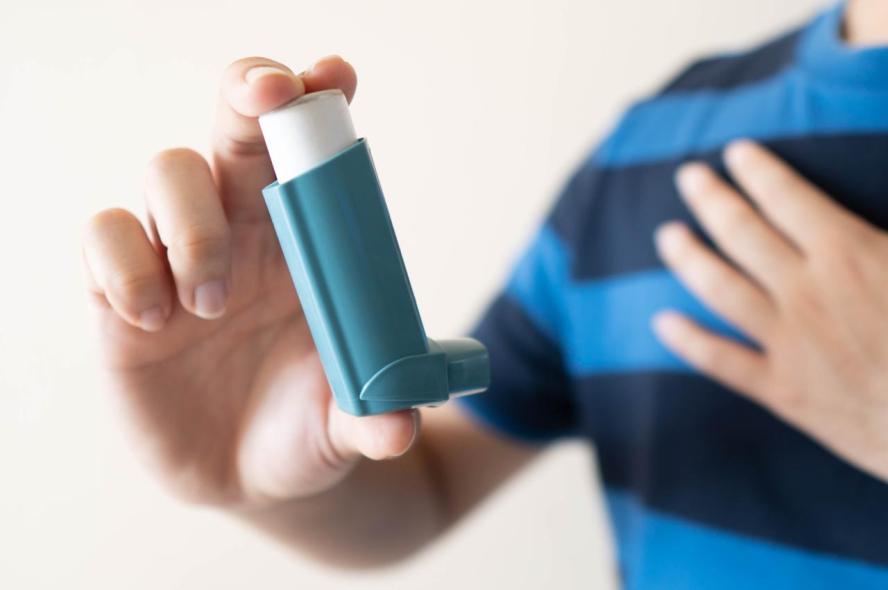 A man holds one hand against his chest and the other outstretched with an inhaler between his index and middle fingers, and his thumb.