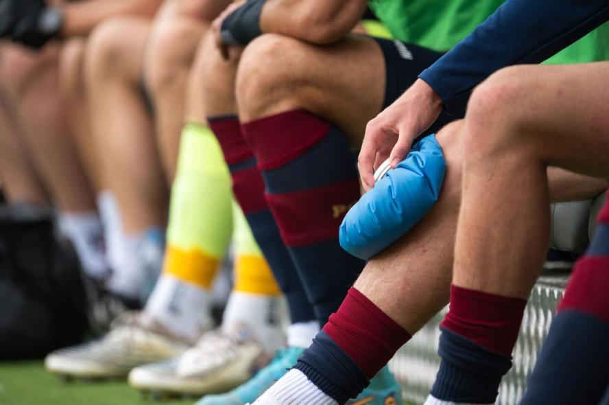 A man holds an ice pack on his knee while seated on a bench alongside other athletes.
