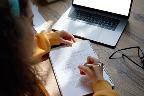 Young woman writing in spiral notebook, with glasses and laptop nearby