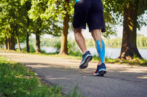 A man's lower body is shown running on a path with blue kinesiology tape applied to his right calf.