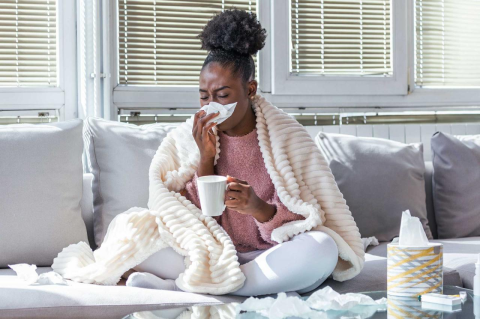 A woman with the flu sits on a gray couch with a cup of tea, wrapped in a soft blanket.