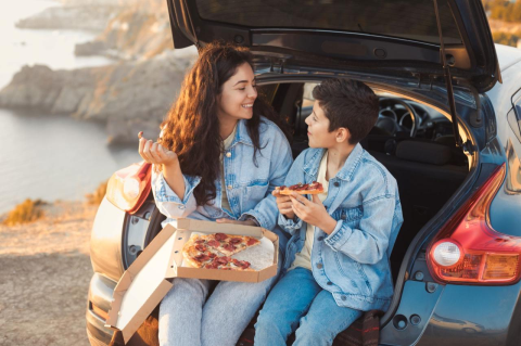 A mother and her teenage son share a pizza dinner on the go, sitting in the open trunk of a small hatchback car. The mother smiles at her talking son, and they are both wearing denim jackets and jeans. 