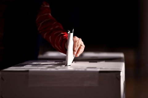 A hand puts a ballot into a ballot box, against a dark background